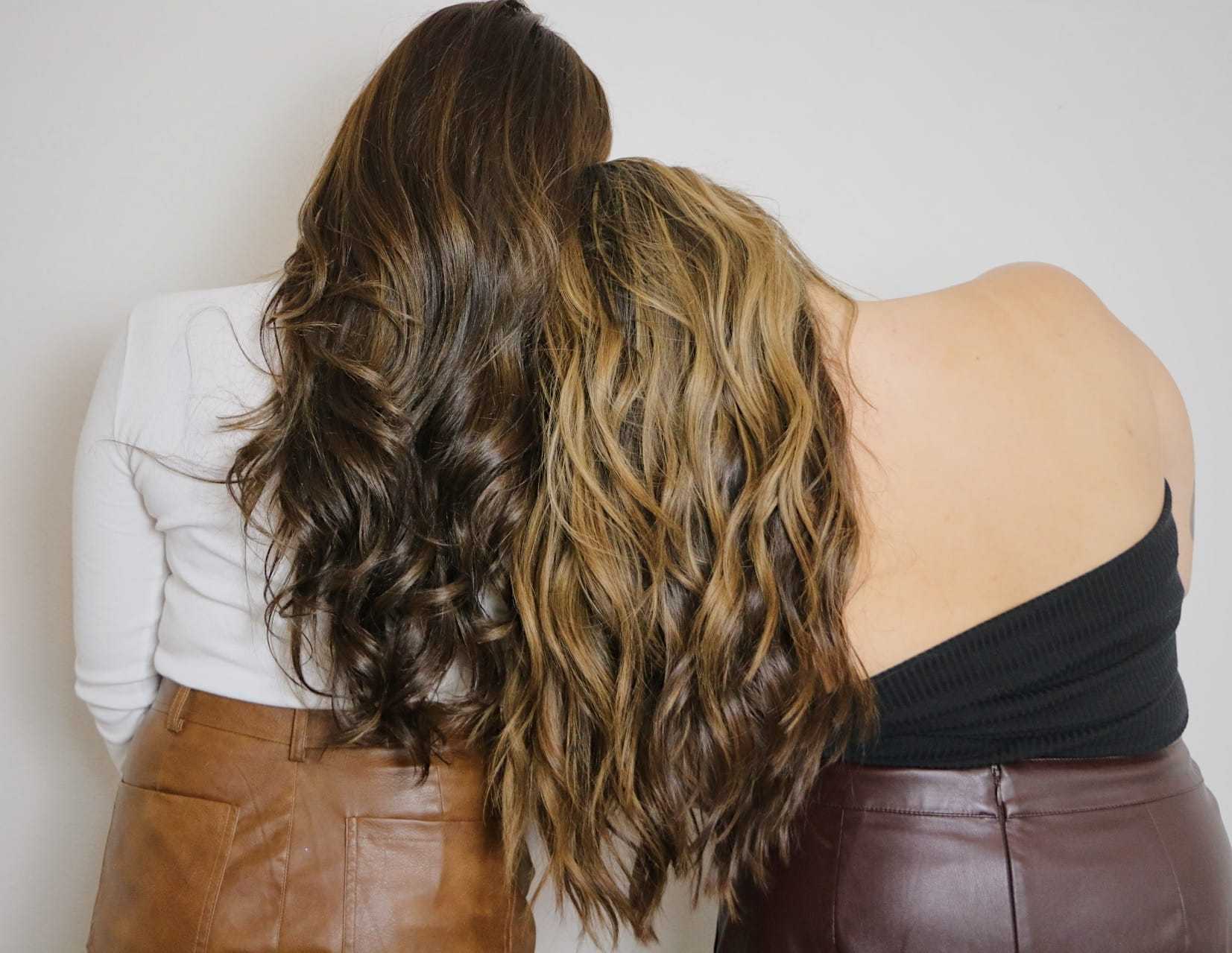 Two women with long, wavy brown hair stand back-to-back against a white background.