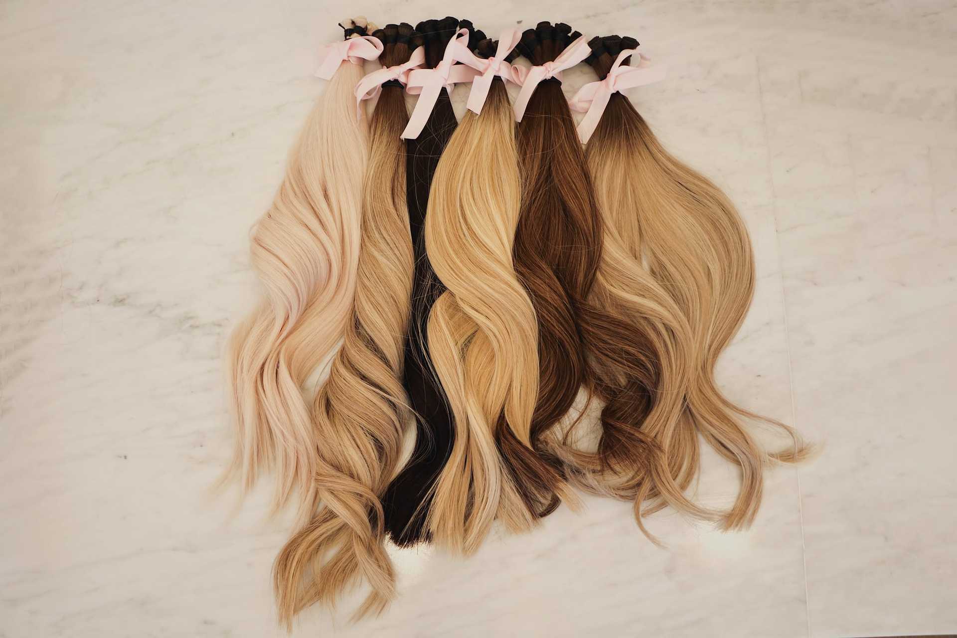 Different shades of blonde and brown hair extensions with pink ribbons on a marble surface.