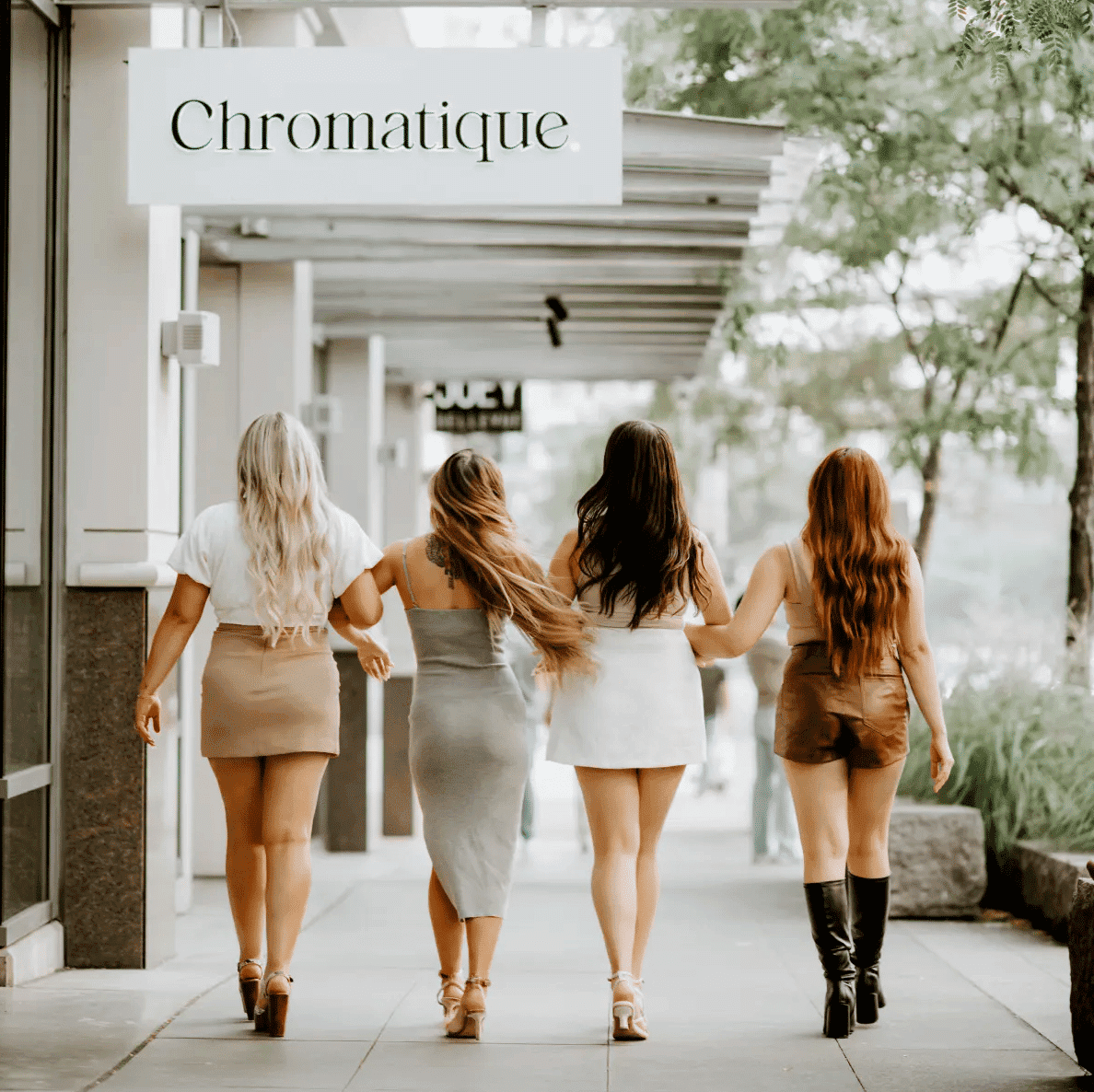 Four women walking arm-in-arm on a city sidewalk under a shop sign.